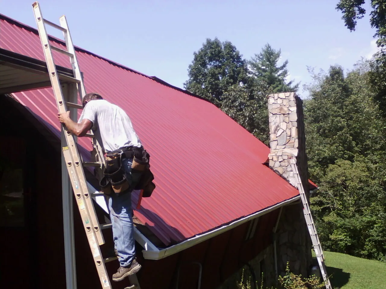 Roofer on a ladder beside a red metal roof and stone chimney — Soffit & Fascia Repair by Roofing Brothers in Old Saybrook